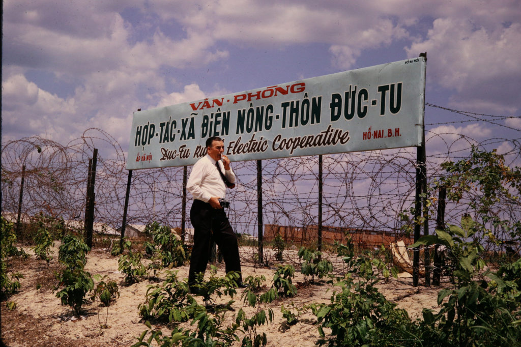 Hugh Bush, NRECA team engineer, stands outside the barbed wire headquarters of Duc Tu Electric Co-op in Ho Nai in 1970. Landmines are hidden in the adjoined field. (Photo Courtesy of Ted Case)